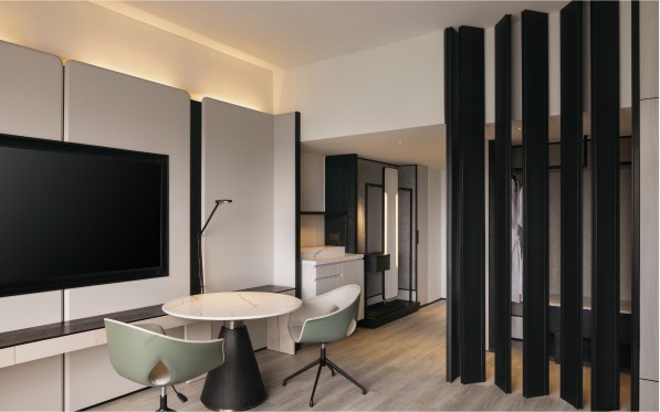 A modern, minimalist interior features a rounded table with chairs, a mounted TV, and sleek dividers creating subtle room separations in a well-lit space.