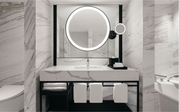 https://hiltonwedding.mediatropy.com/wp-content/uploads/2024/01/bridal6.jpg A modern bathroom vanity with a circular, illuminated mirror and a marble countertop, set against marble tiled walls. The space includes a faucet, towels, and a reflection of a toilet.