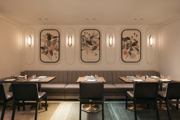 A modern dining area features a long bench with tables set for a meal, framed botanical prints on the wall, and warm lighting. The ambiance is elegant and inviting.