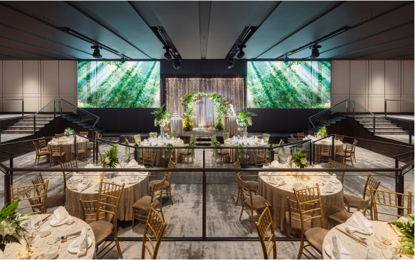 Elegantly set round tables with chairs fill a venue hall, facing a large stage with greenery-themed backdrops for an event, potentially a wedding.