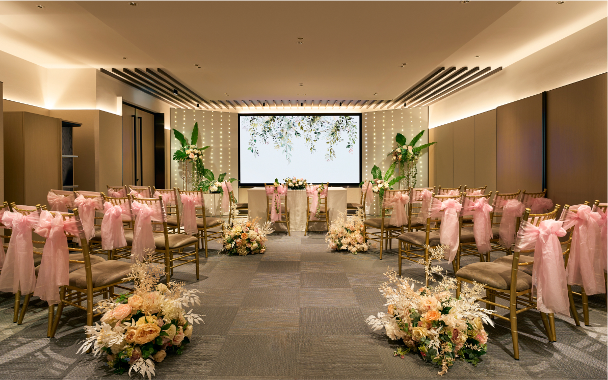 Rows of gold chairs with pink sashes line a carpeted aisle leading to a screen with a floral image, flanked by flower arrangements and lighted pillars, in an elegant indoor setting.