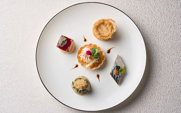 https://hiltonwedding.mediatropy.com/wp-content/uploads/2024/01/the-manor-13.jpg Elegant appetizers arranged artistically on a large white plate with a black rim, against a textured light background.